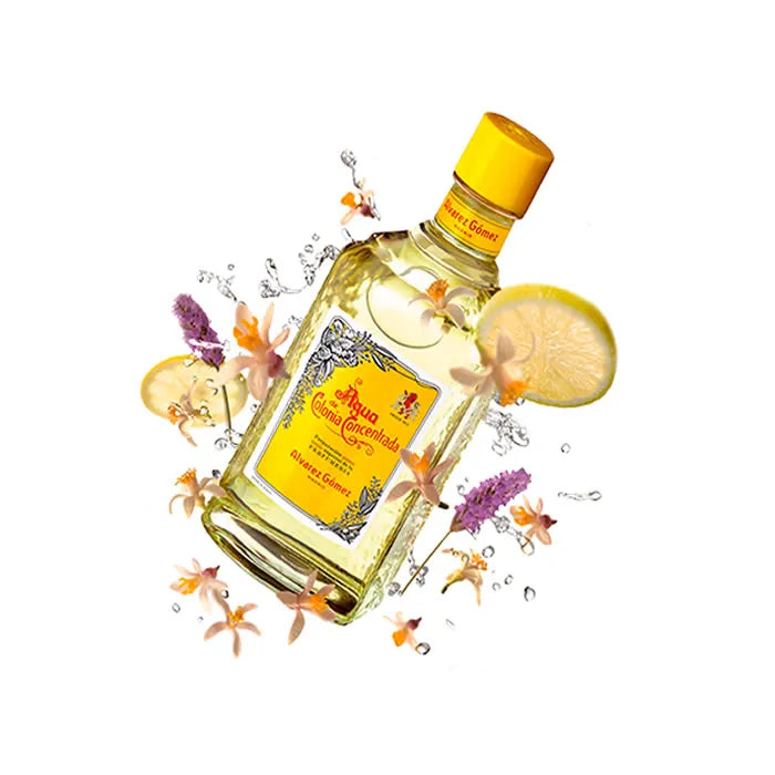 Bottiglia di acqua di colonia concentrata di colore giallo di Alvarez Gomez, inclinata, con fiori, fette di limone e schizzi d'acqua su sfondo bianco.