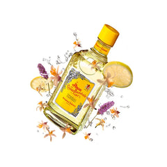 Bottiglia di acqua di colonia concentrata di colore giallo di Alvarez Gomez, inclinata, con fiori, fette di limone e schizzi d'acqua su sfondo bianco.